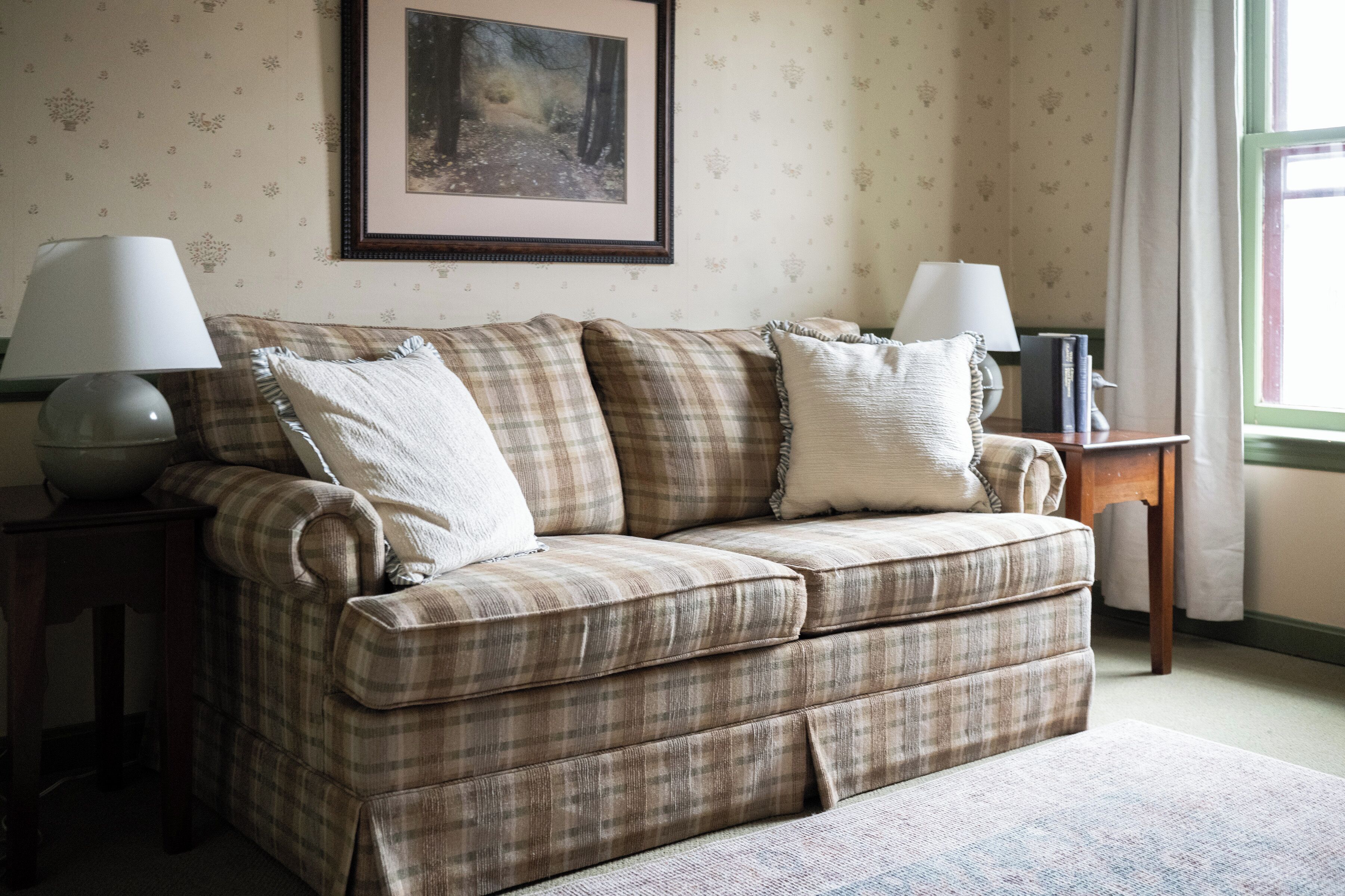 A living room with a plaid sofa, two lamps, a framed picture on the wall, and a window with curtains.