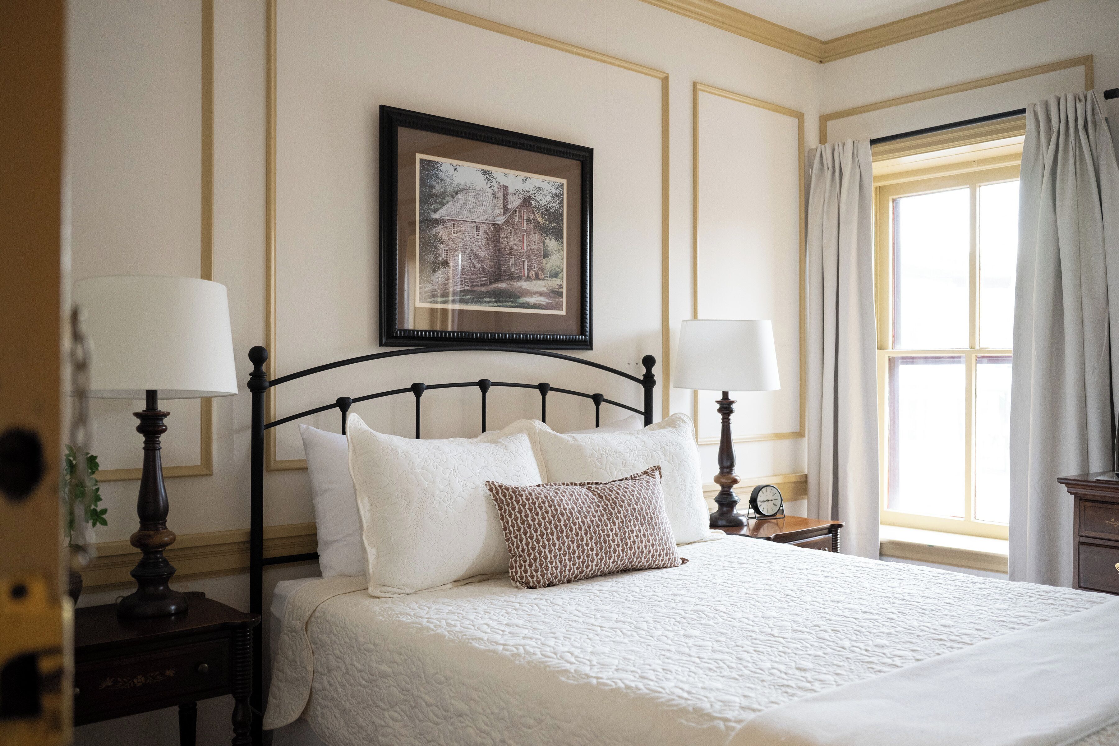 A neatly made bed with a headboard, two bedside tables with lamps, a framed picture on the wall, and a window with curtains.