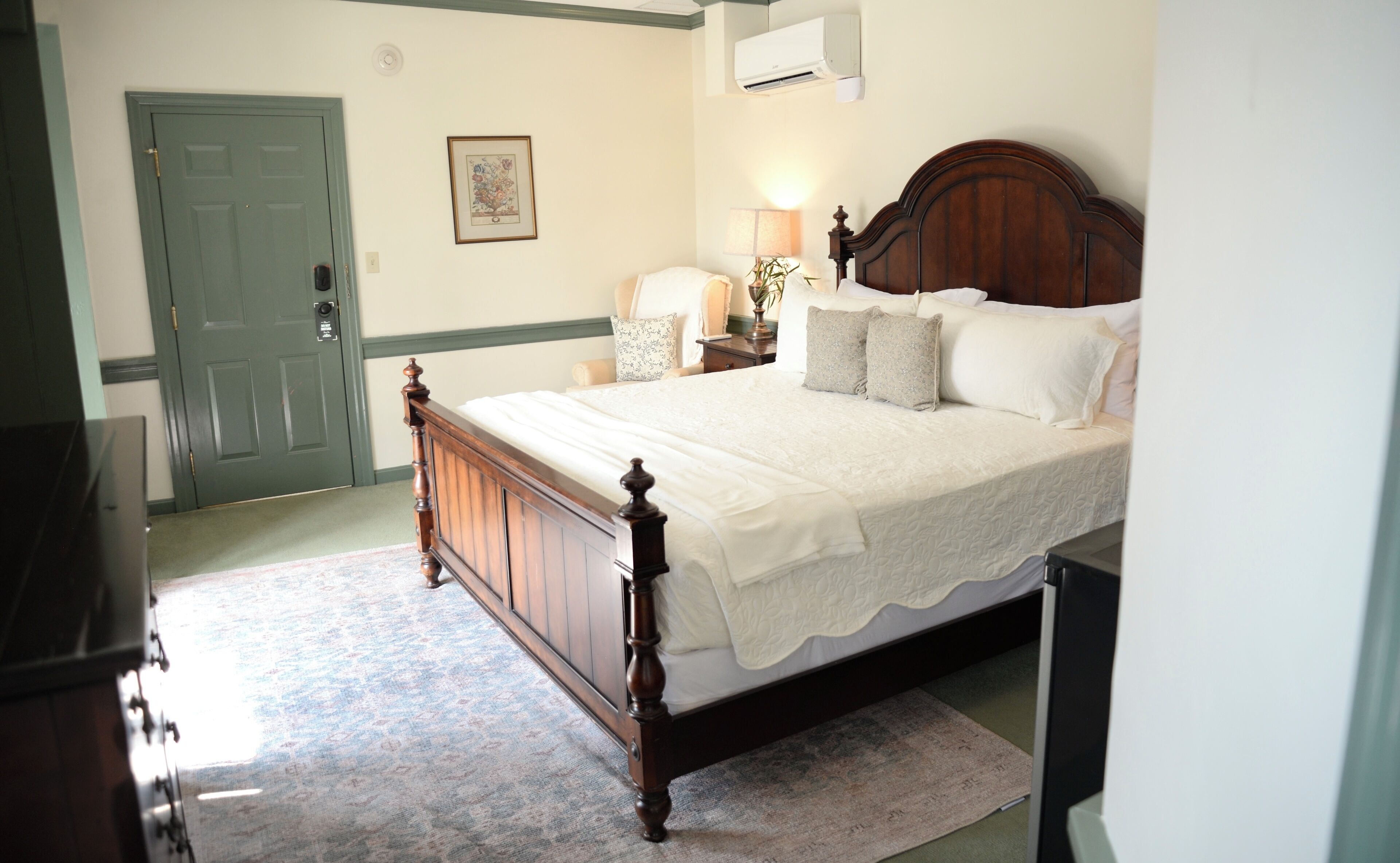 A bedroom with a wooden bed, a green door, a framed picture on the wall, and a nightstand with a lamp.