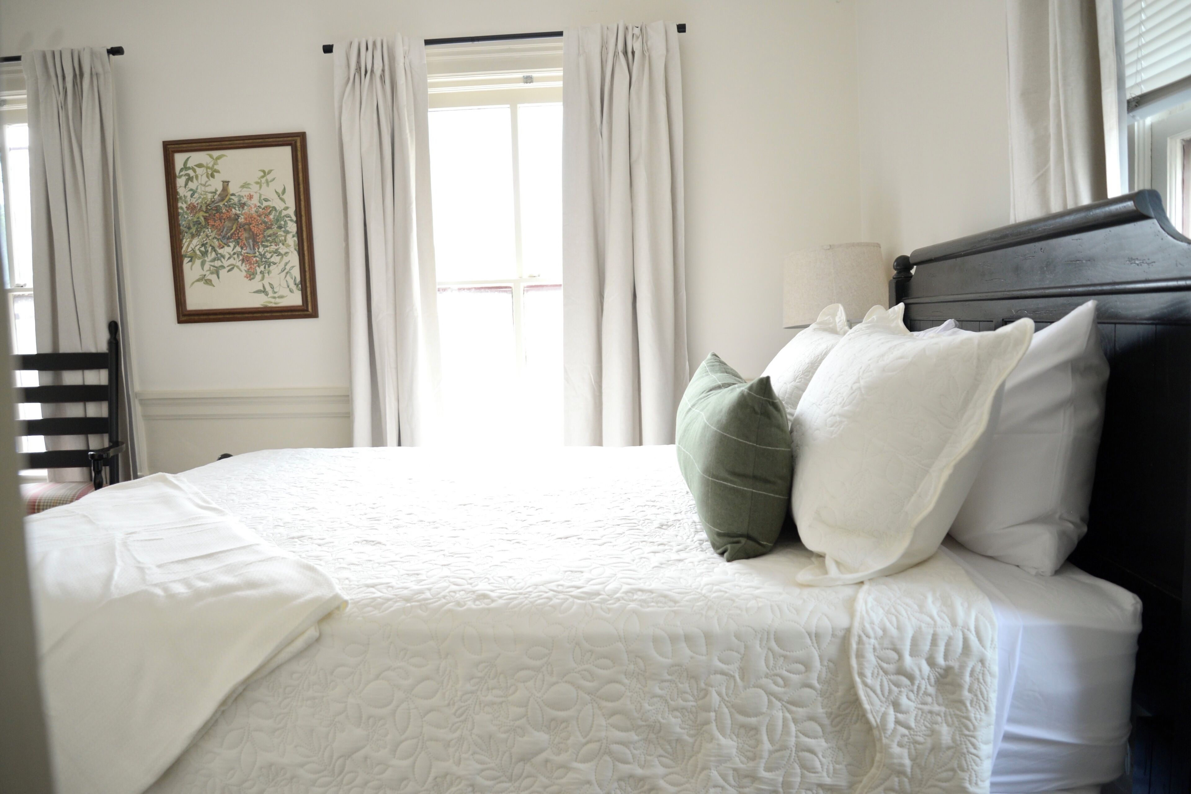 A neatly made bed with white bedding and a dark headboard, a framed floral artwork on the wall, and a window with curtains.