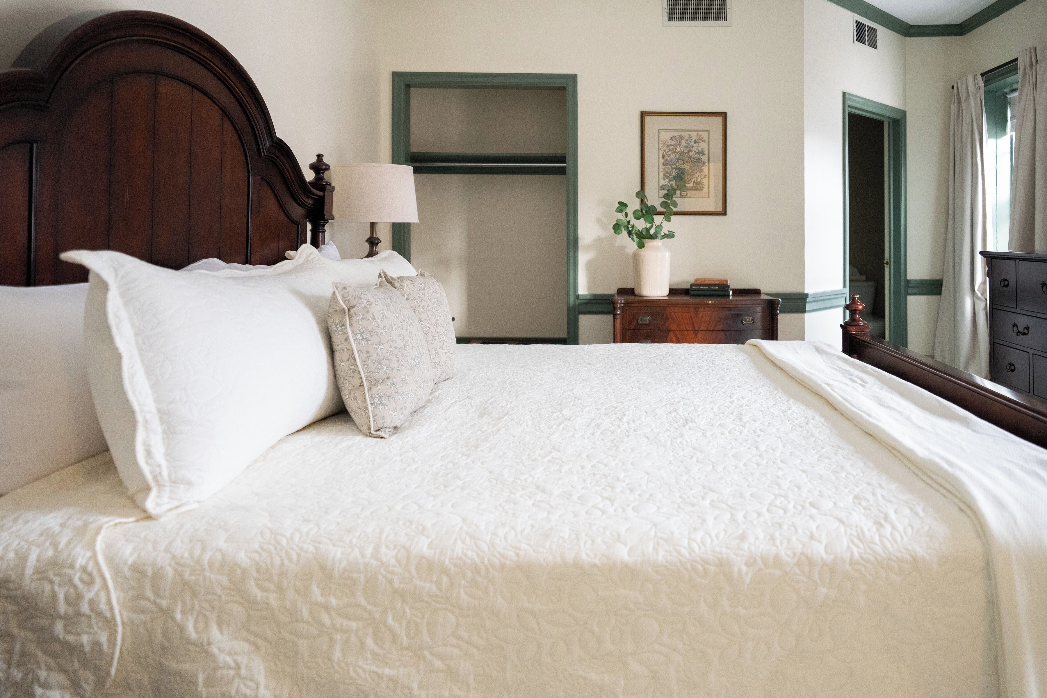 A neatly made bed with a white bedspread and pillows, a wooden headboard, a bedside table with a lamp, a dresser, and a framed picture on the wall.