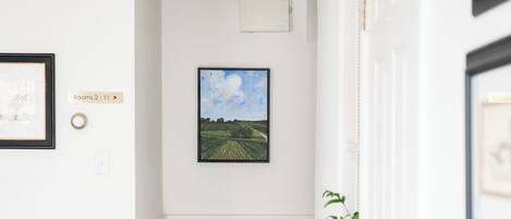 Un couloir avec une porte blanche, un tableau représentant un paysage encadré, un banc blanc avec un coussin bleu et un tapis à motifs.
