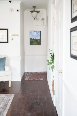 Un pasillo con una puerta blanca, un cuadro de paisaje enmarcado, un banco blanco con cojín azul y una alfombra estampada.