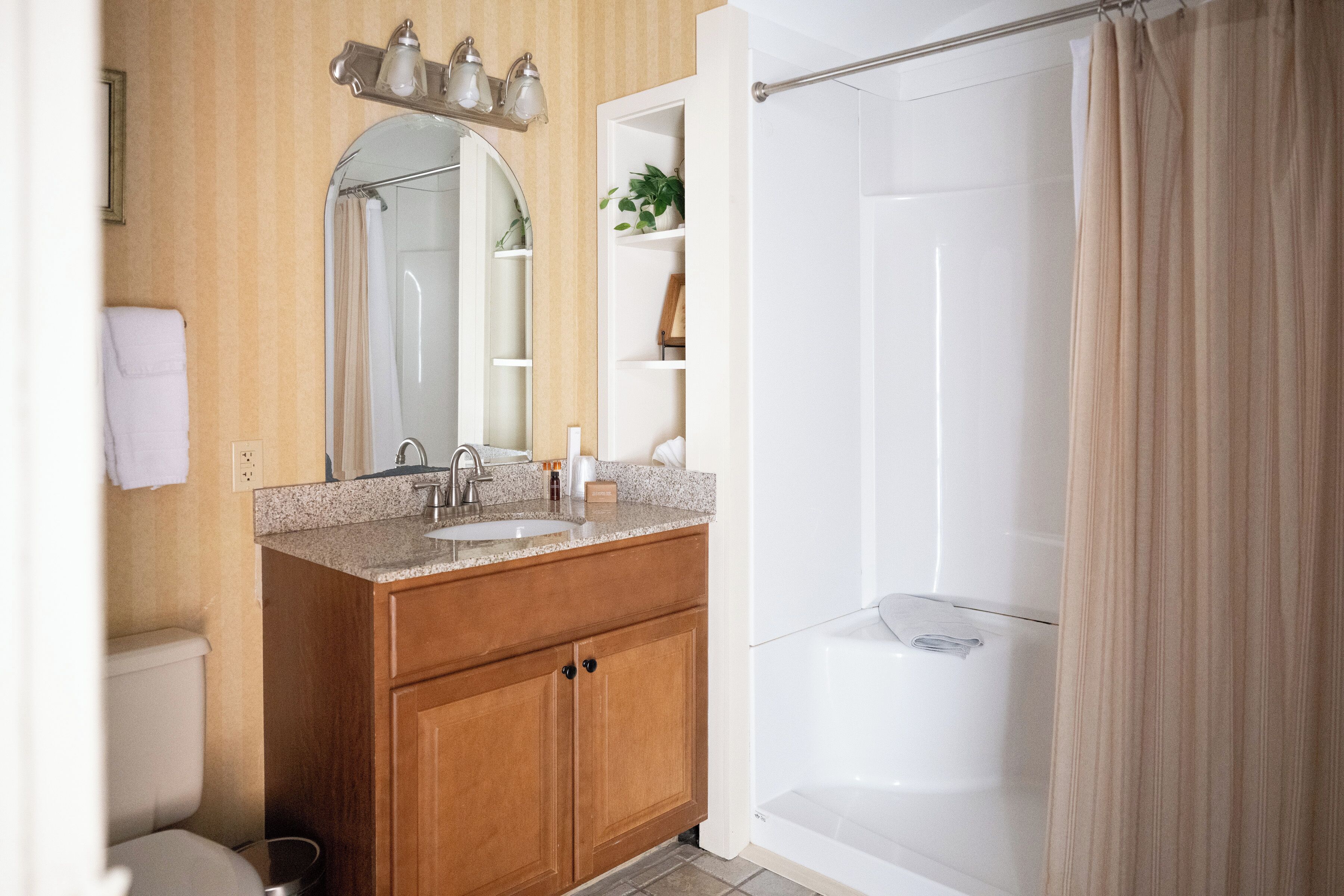 A bathroom with a wooden vanity, a sink, a mirror, a shower with a curtain, and a shelf with toiletries.