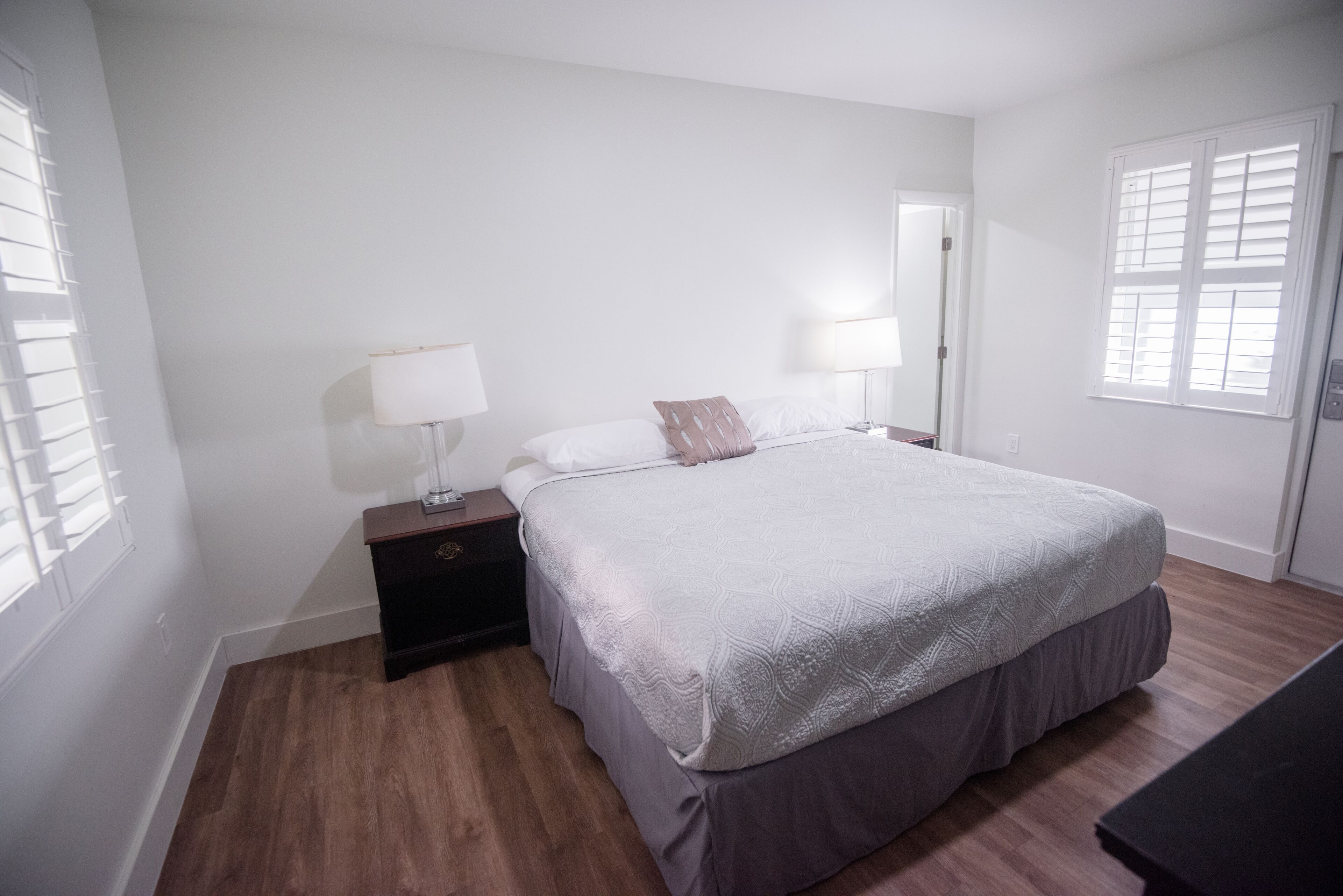 Un dormitorio con una cama grande, dos mesitas de noche con lámparas, piso de madera y paredes blancas.