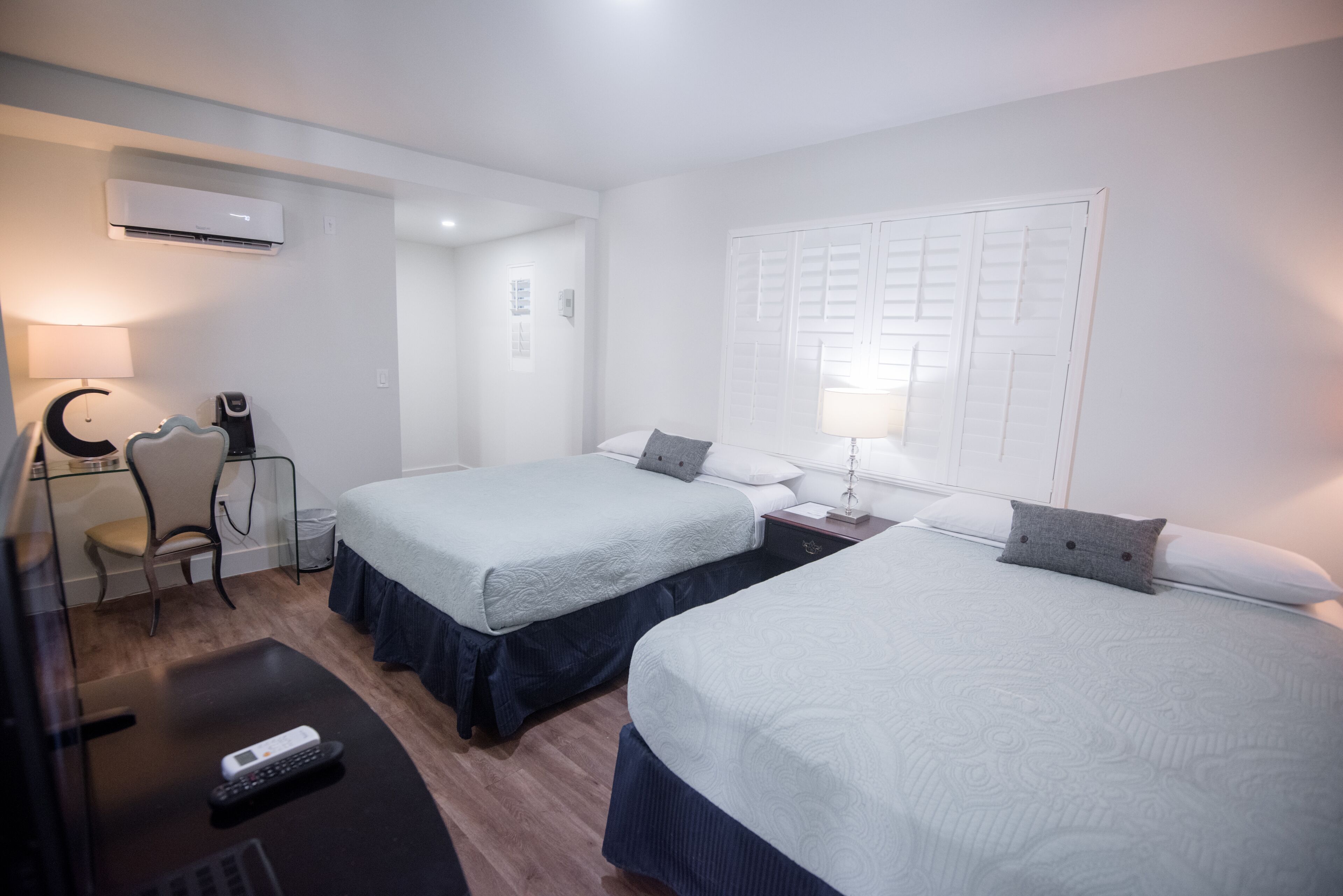 A hotel room with two beds, a desk with a chair, a lamp, and an air conditioning unit.