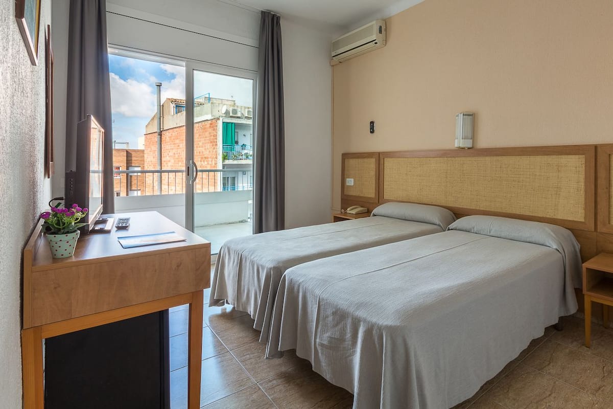 A hotel room with two beds, a desk, and a view of buildings through a large window.