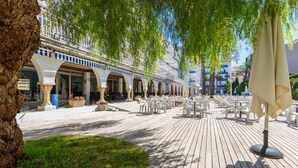 An outdoor patio area with white tables and chairs, surrounded by trees and a building with balconies. - Hotel Apartamentos Solimar (Calafell)