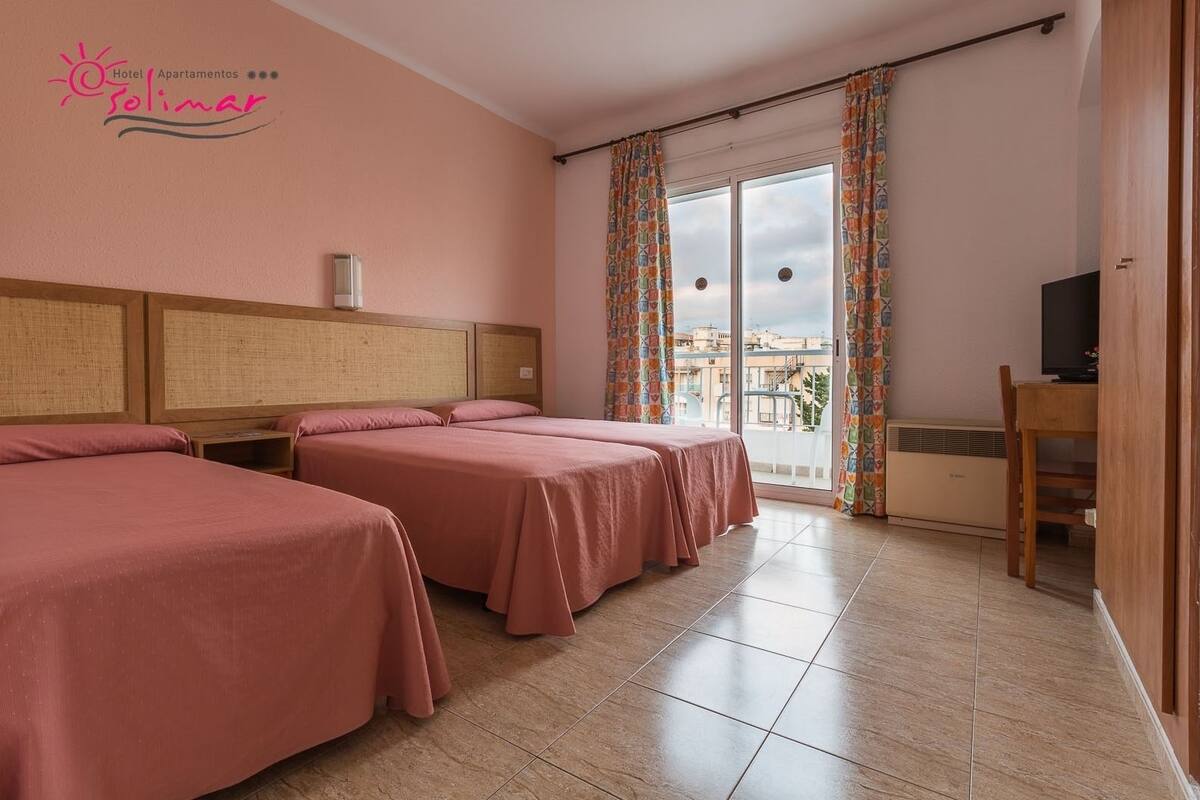 A hotel room with two beds, a TV, a desk, and a balcony with curtains.