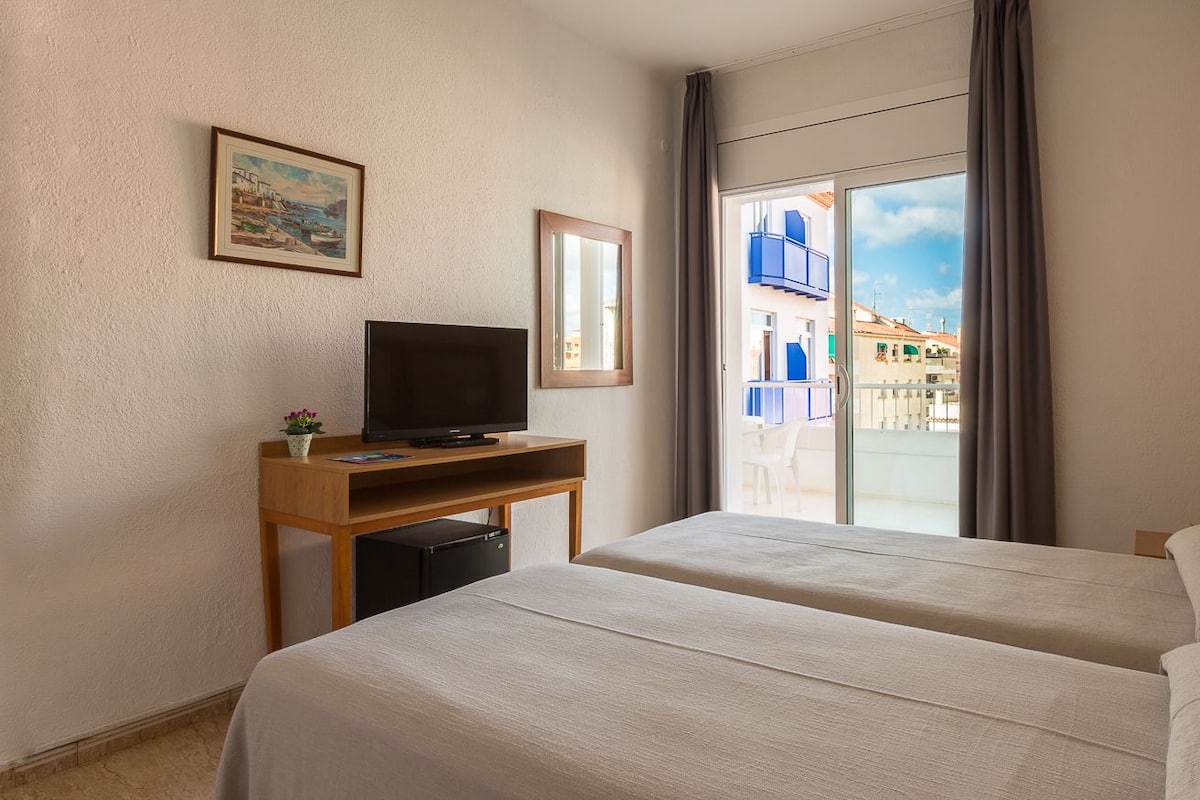 A hotel room with two beds, a TV on a wooden stand, a small table with a plant, and a view of buildings through a glass door.