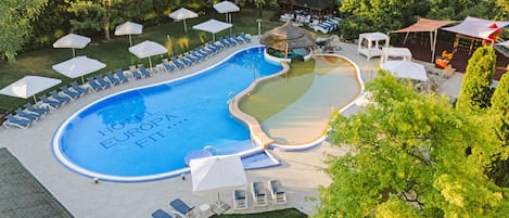 Una piscina con sillas de descanso y sombrillas, rodeada de árboles y un edificio pequeño.