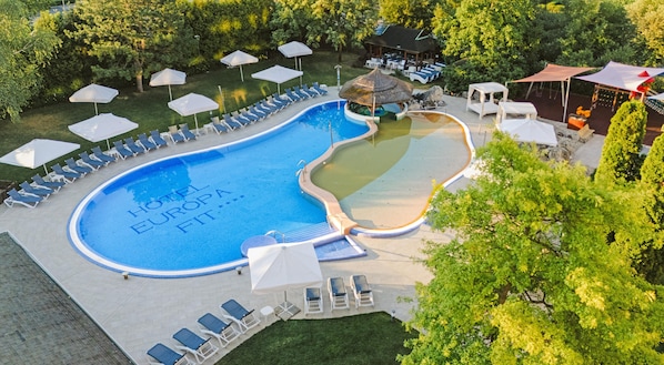 Una piscina con sillas de descanso y sombrillas, rodeada de árboles y un edificio pequeño.
