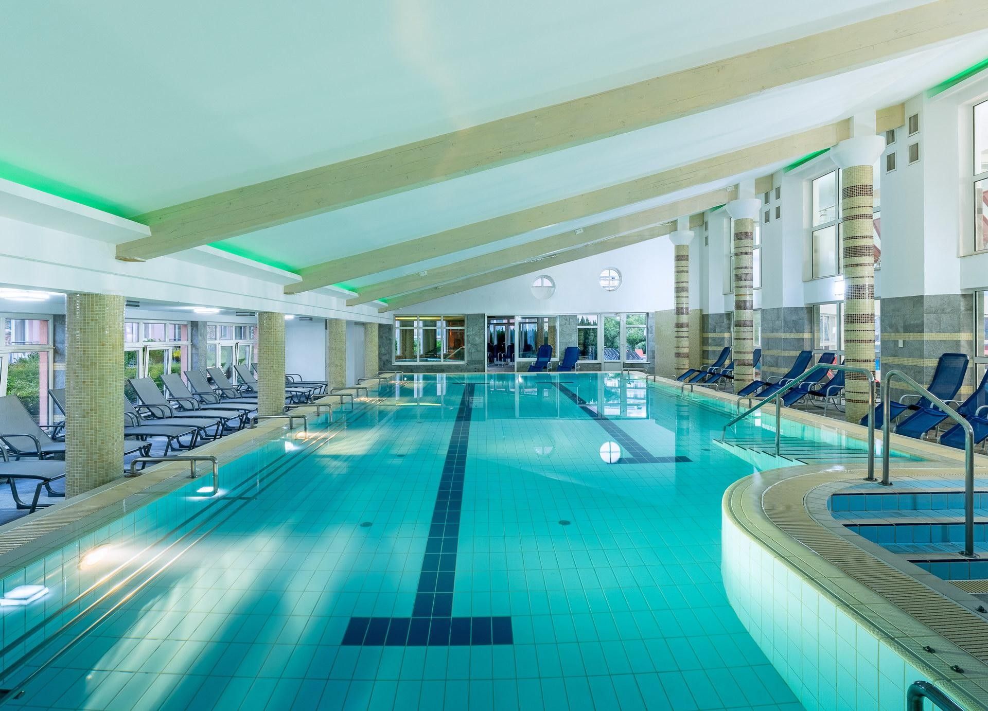 Ein großes Hallenbad mit mehreren Bahnen, Liegestühlen und Blick ins Freie.