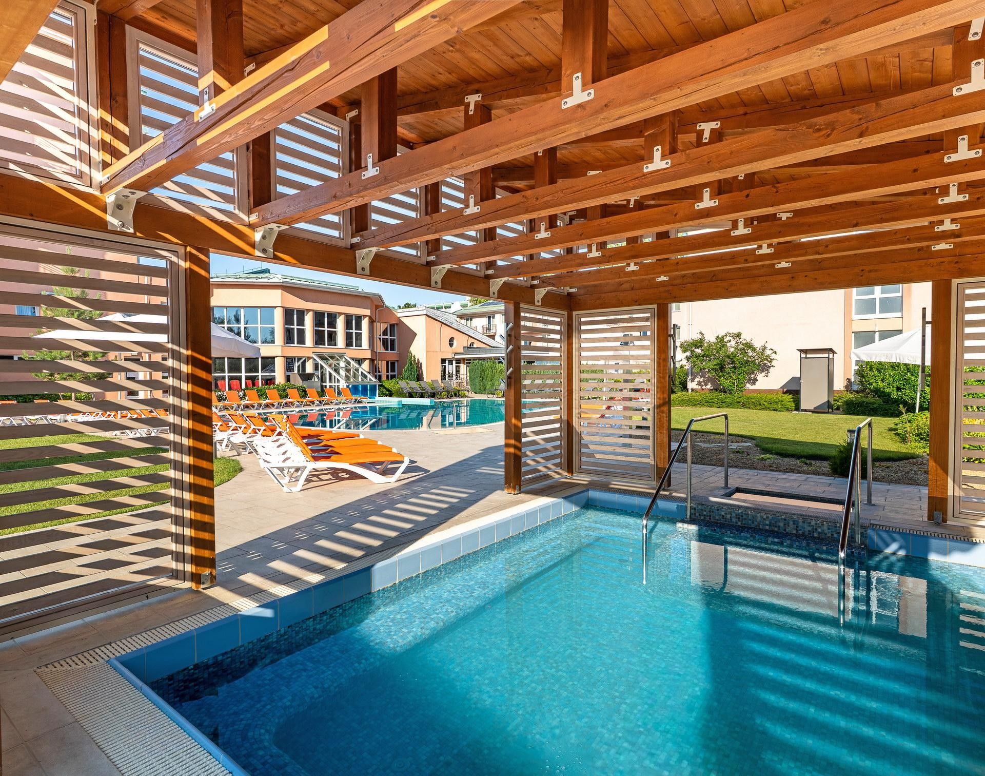 A swimming pool with a wooden deck and lounge chairs, surrounded by buildings.
