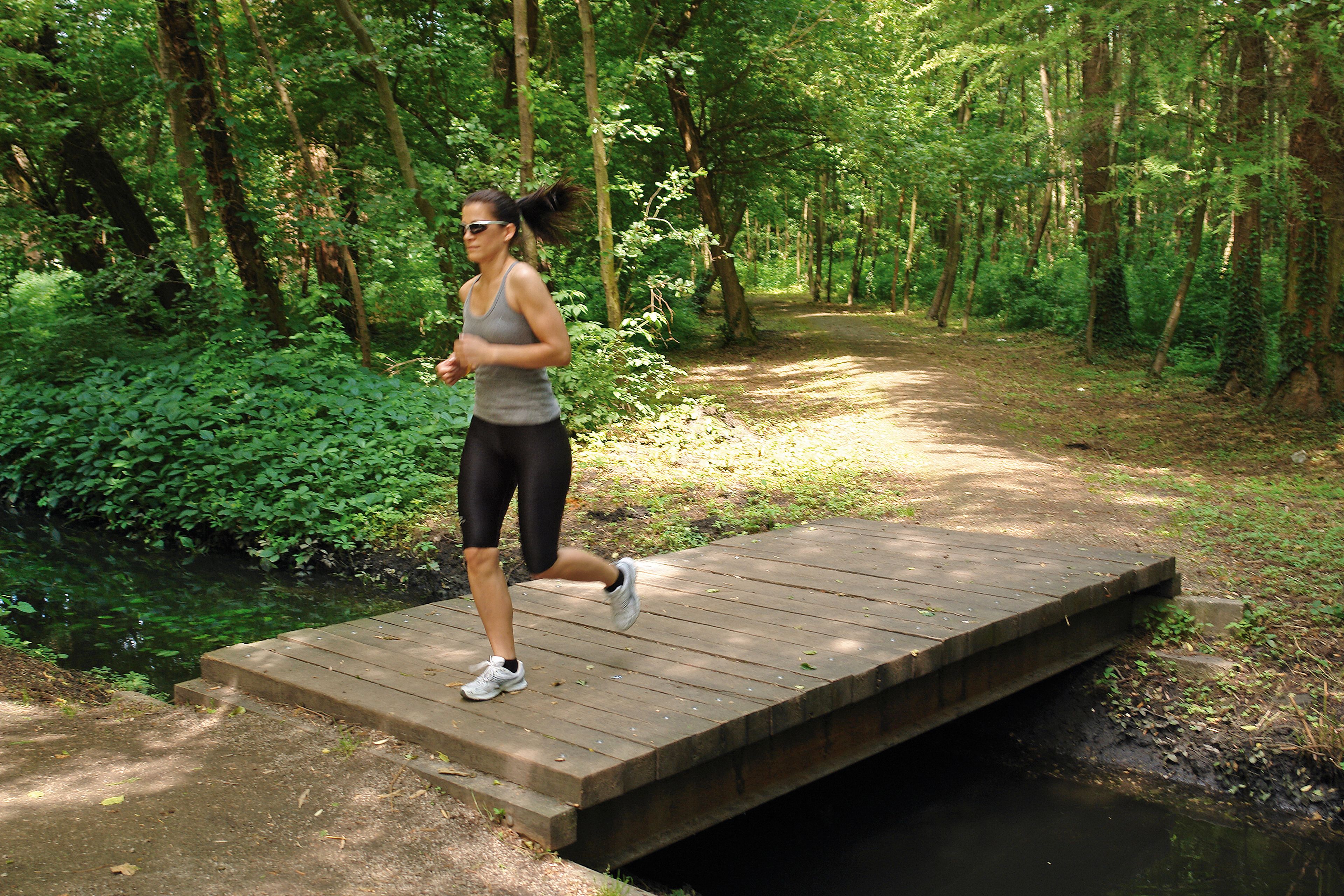 A woman jogging on a wooden bridge over a small stream in a forested area.