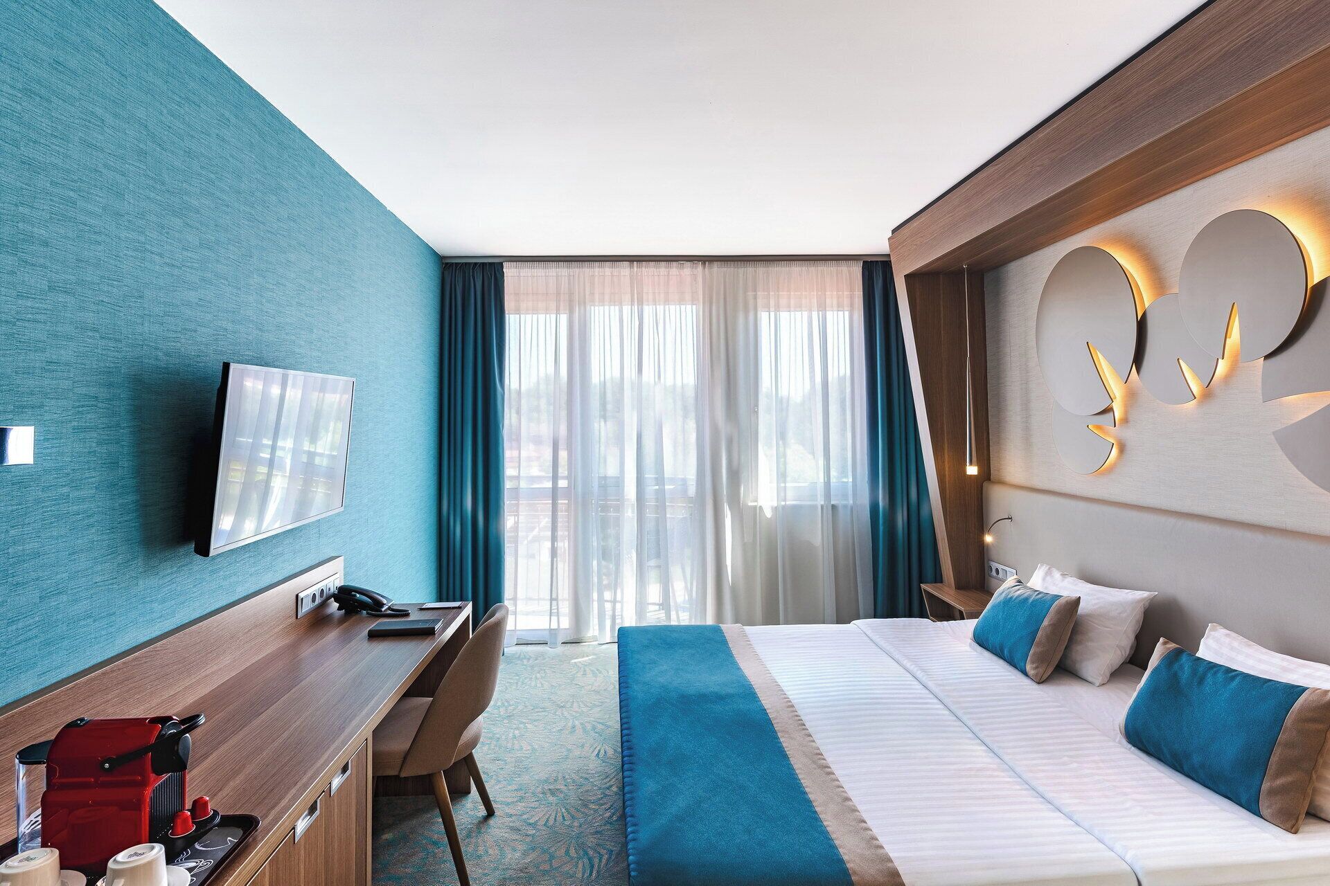 A hotel room with a bed, a desk, a coffee machine, a TV, and a large window with curtains.