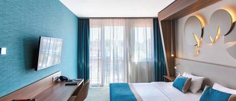 A hotel room with a bed, a desk, a coffee machine, a TV, and a large window with curtains.