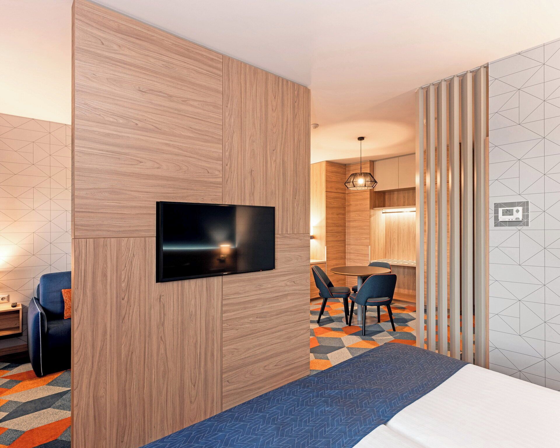 A hotel room with a flat-screen TV mounted on a wooden panel, a dining area with a table and chairs, and a patterned carpet.