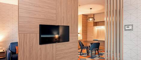 A hotel room with a flat-screen TV mounted on a wooden panel, a dining area with a table and chairs, and a patterned carpet.