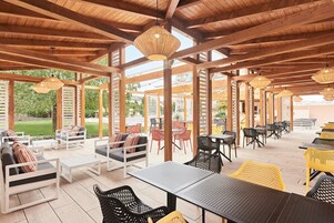 Eine Terrasse im Freien mit Holzbalken, Tischen, Stühlen und hängenden Lichtern.