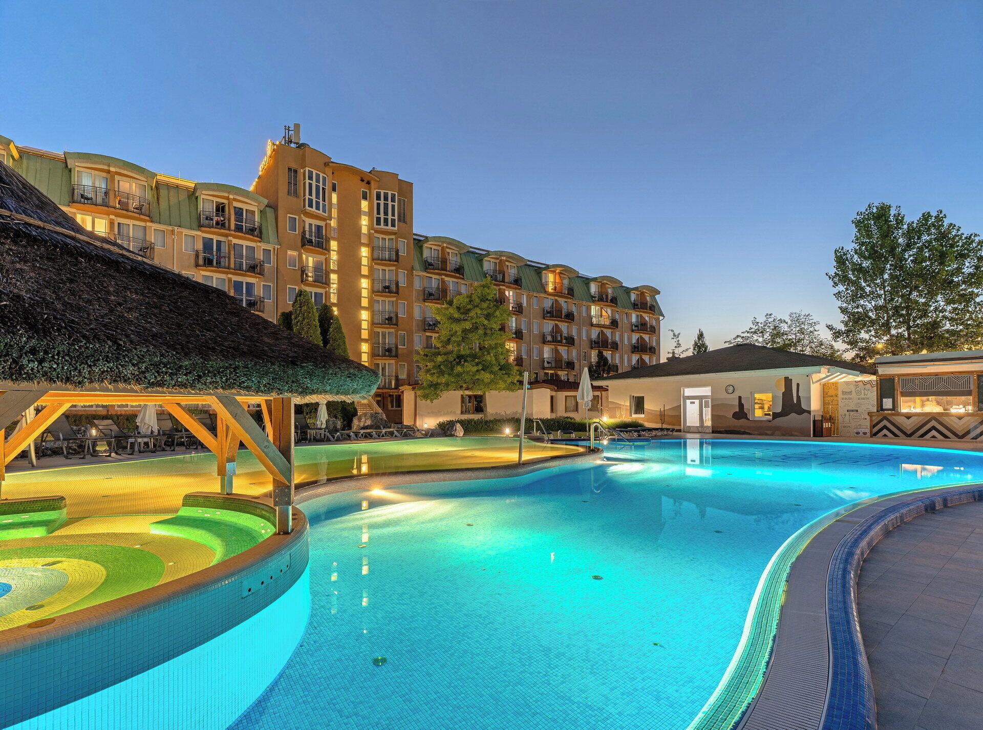 Une piscine dotée d’une zone plus profonde, entourée d’un bâtiment comportant de nombreux balcons et surmontée d’une structure à toit de chaume.