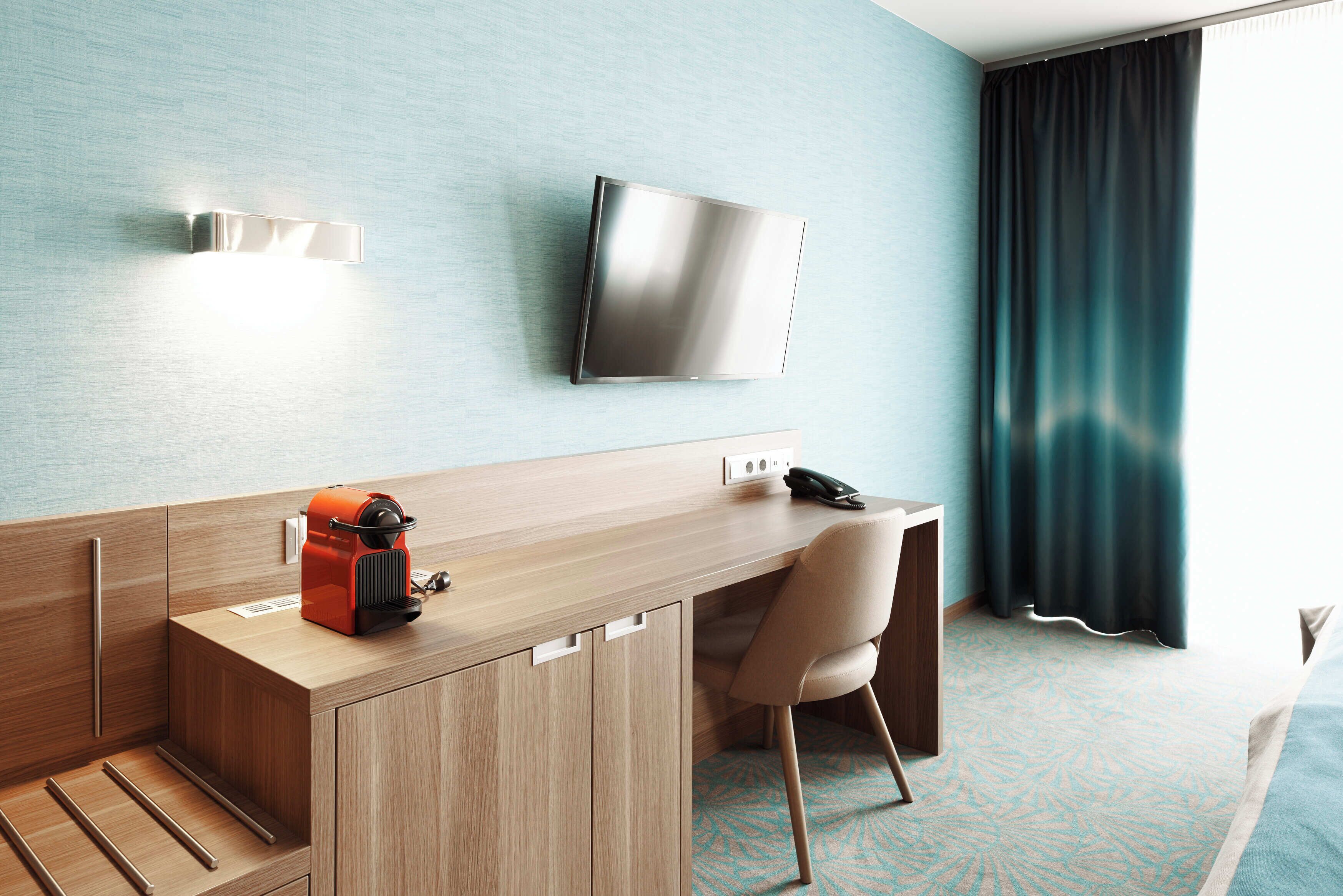 A hotel room with a wooden desk, a chair, a flat-screen TV mounted on the wall, and a window with curtains.