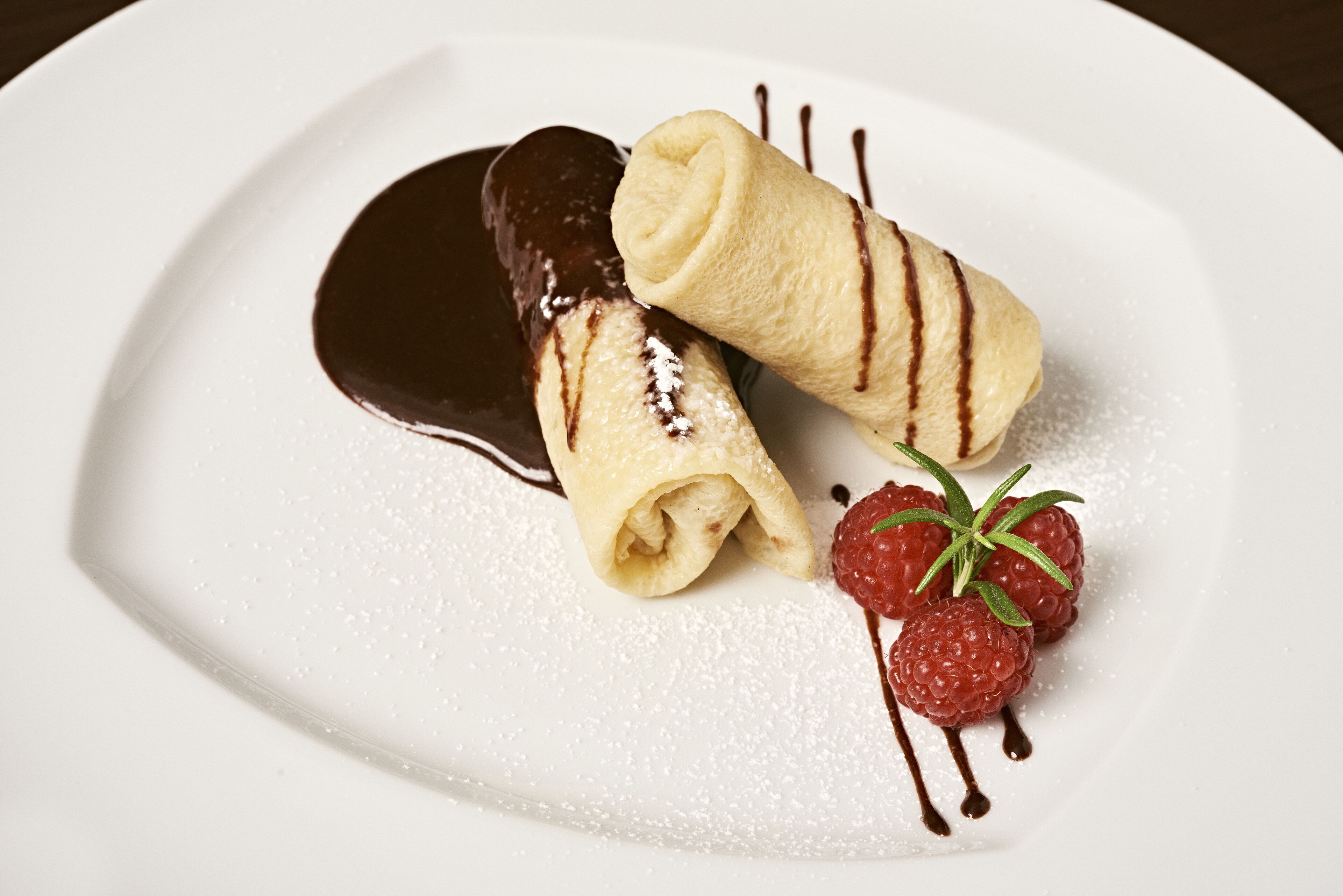 A dessert plate with two rolled crepes, chocolate sauce, powdered sugar, and a raspberry with a sprig of rosemary.