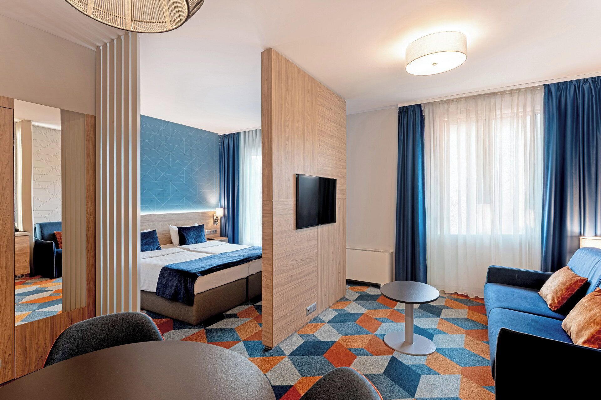 Una habitación de hotel con cama, zona de estar, televisor y un ventanal con cortinas.