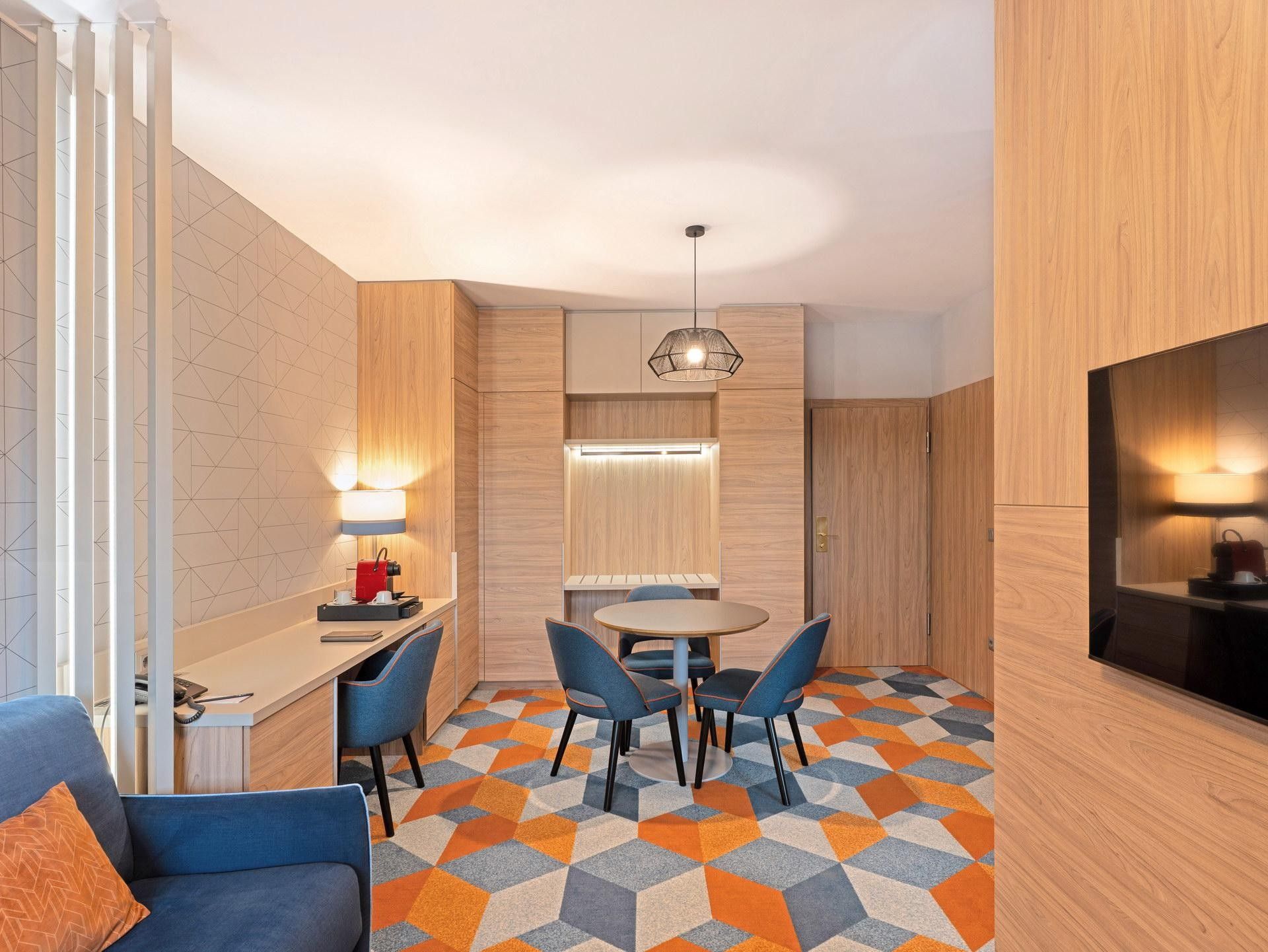 A modern hotel room with a sofa, a dining table with chairs, a TV, and a patterned floor.