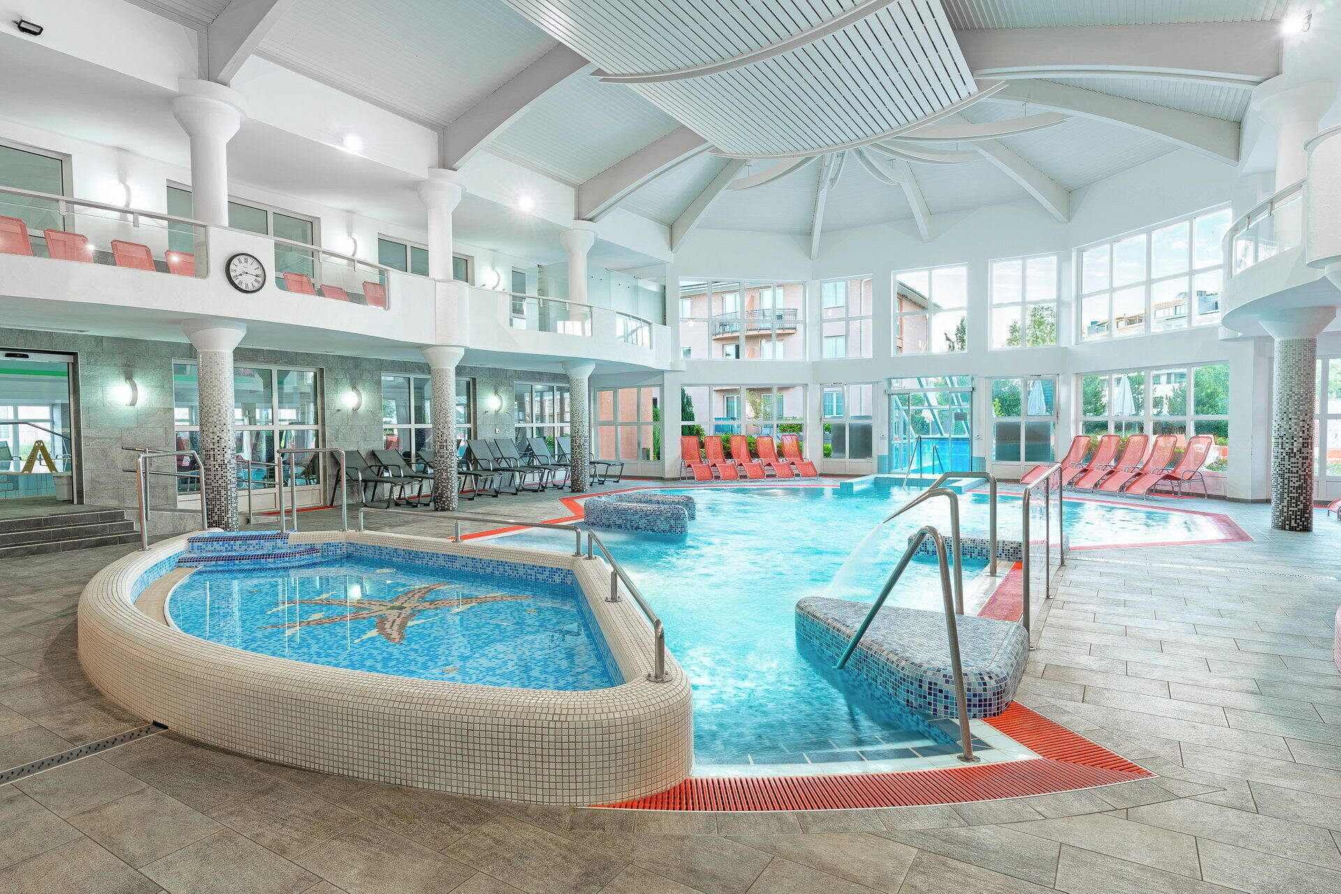 Une grande piscine intérieure dotée de plusieurs couloirs, entourée de chaises longues et surmontée d’un haut plafond laissant entrer la lumière naturelle.