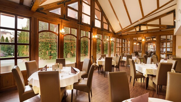 A restaurant with round tables and chairs, large windows with views of greenery, and wooden beams on the ceiling. - Hotel Európa Fit (Hévíz)