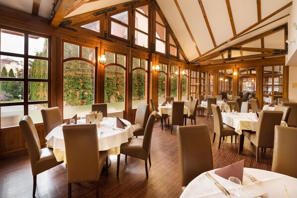A restaurant with round tables and chairs, large windows with views of greenery, and wooden beams on the ceiling. - Hotel Európa Fit (Hévíz)