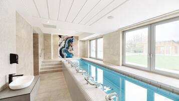 Une piscine intérieure moderne, de forme rectangulaire et allongée, dotée d’un banc intégré le long du bord et offrant une vue sur l’extérieur grâce à de grandes fenêtres.