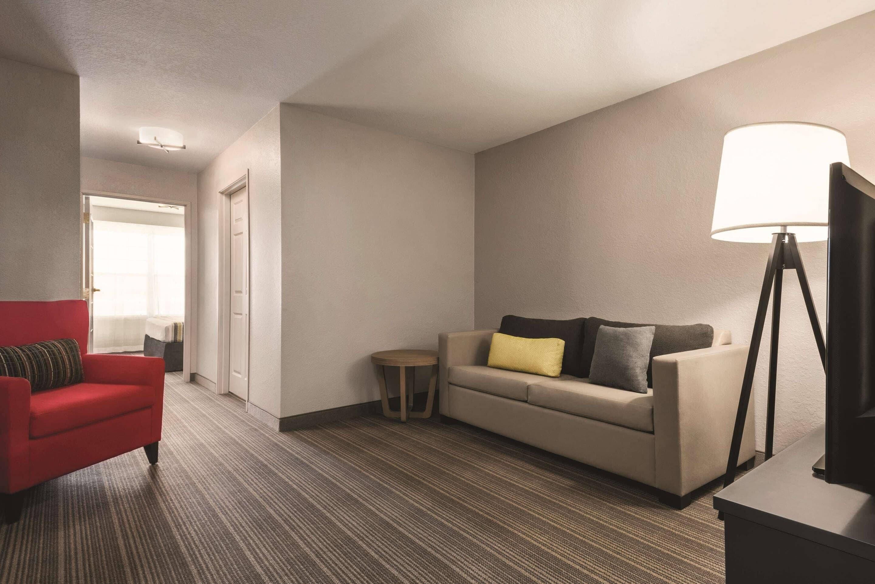A hotel room with a red armchair, a beige sofa with cushions, a small round table, and a floor lamp.