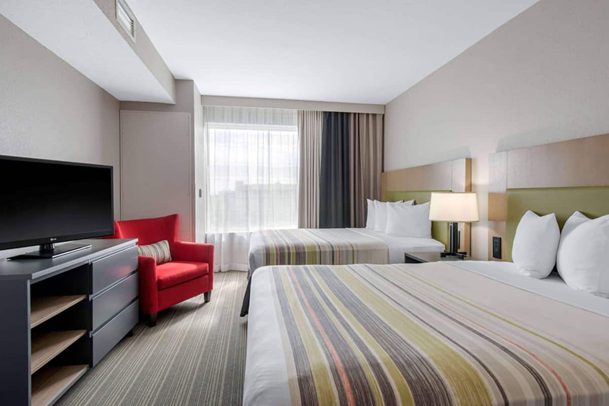 A hotel room with a TV, a red chair, two beds with striped bedding, a lamp, and a large window with curtains.