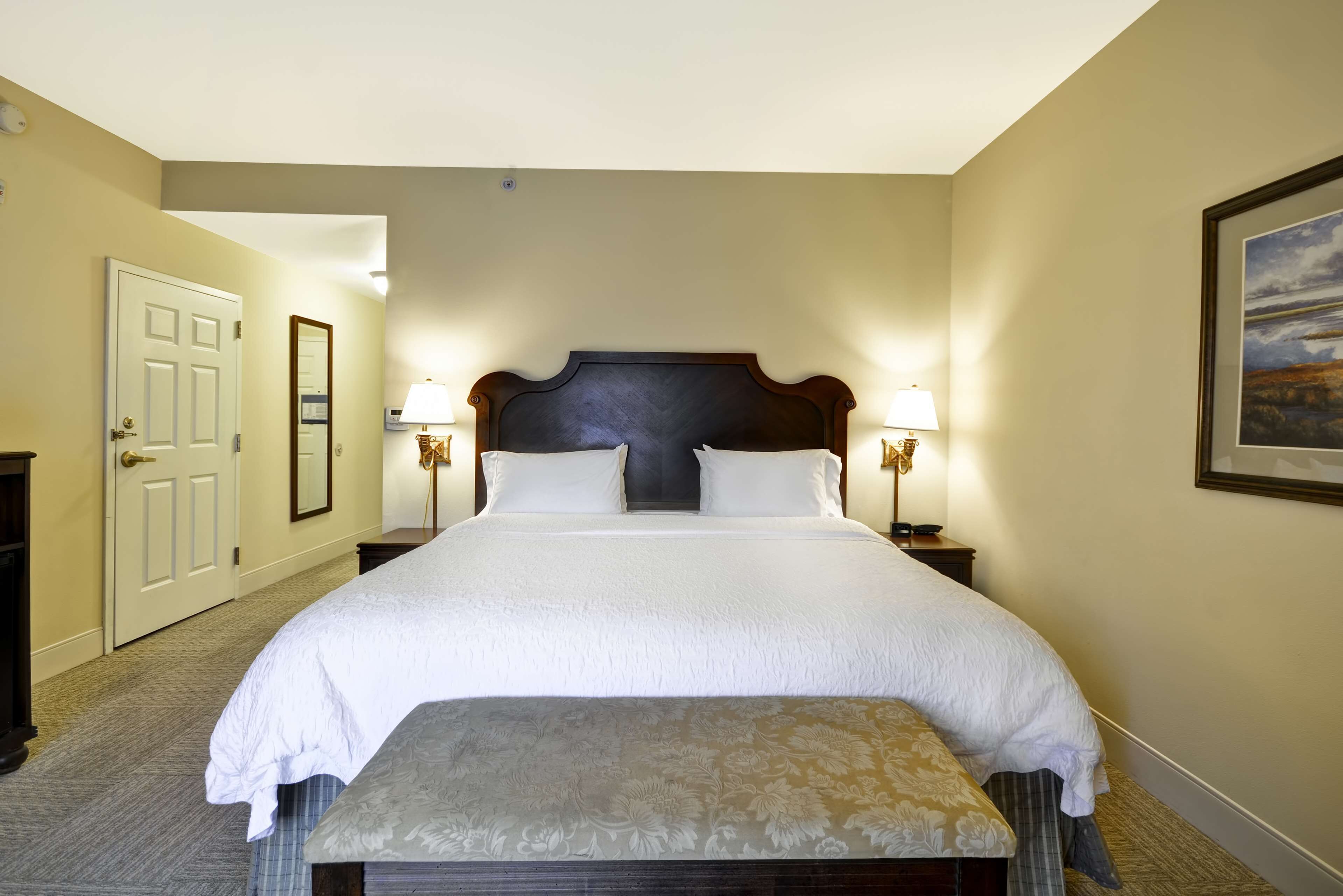 A hotel room with a large bed, two bedside lamps, a door, a mirror, and a framed picture on the wall.