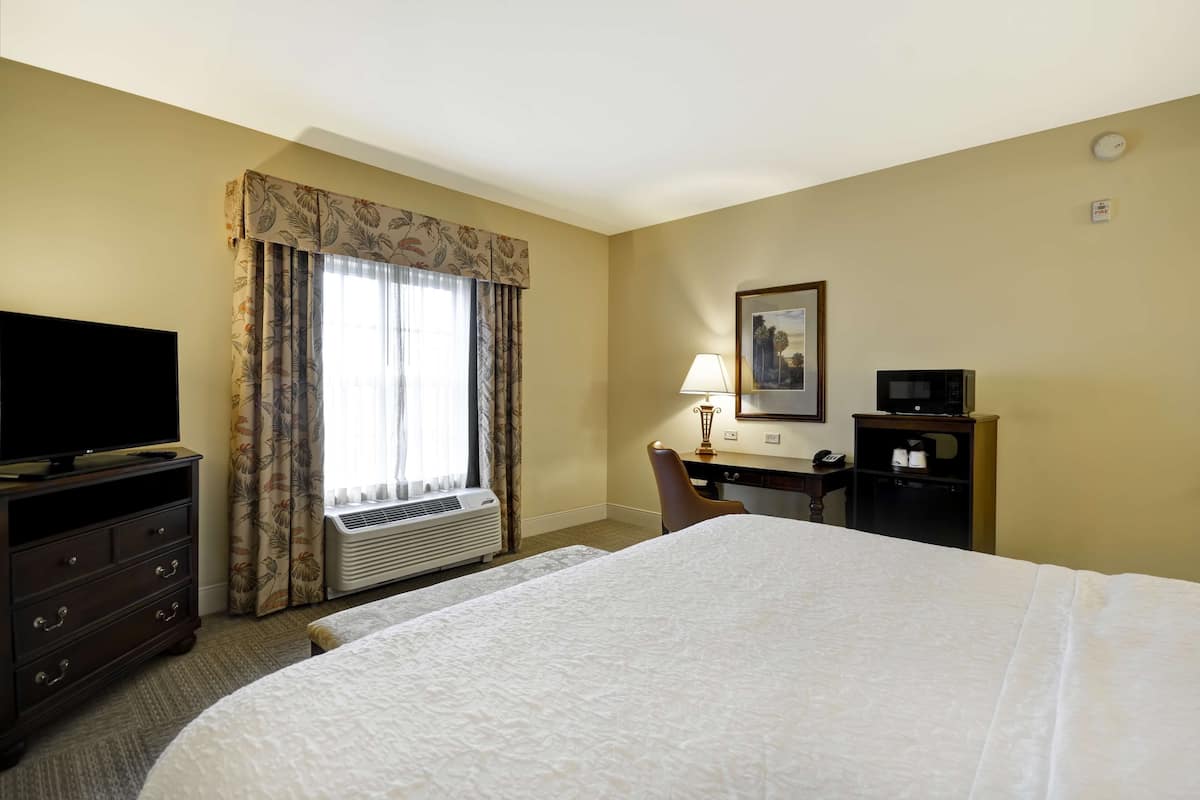 A hotel room with a large bed, a television on a dresser, a desk with a lamp, a chair, and a microwave.