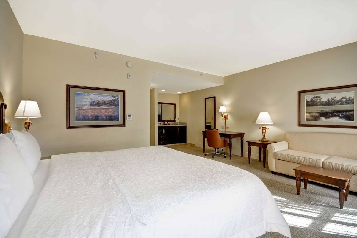A hotel room with a large bed, a desk, a chair, a sofa, and a small table.