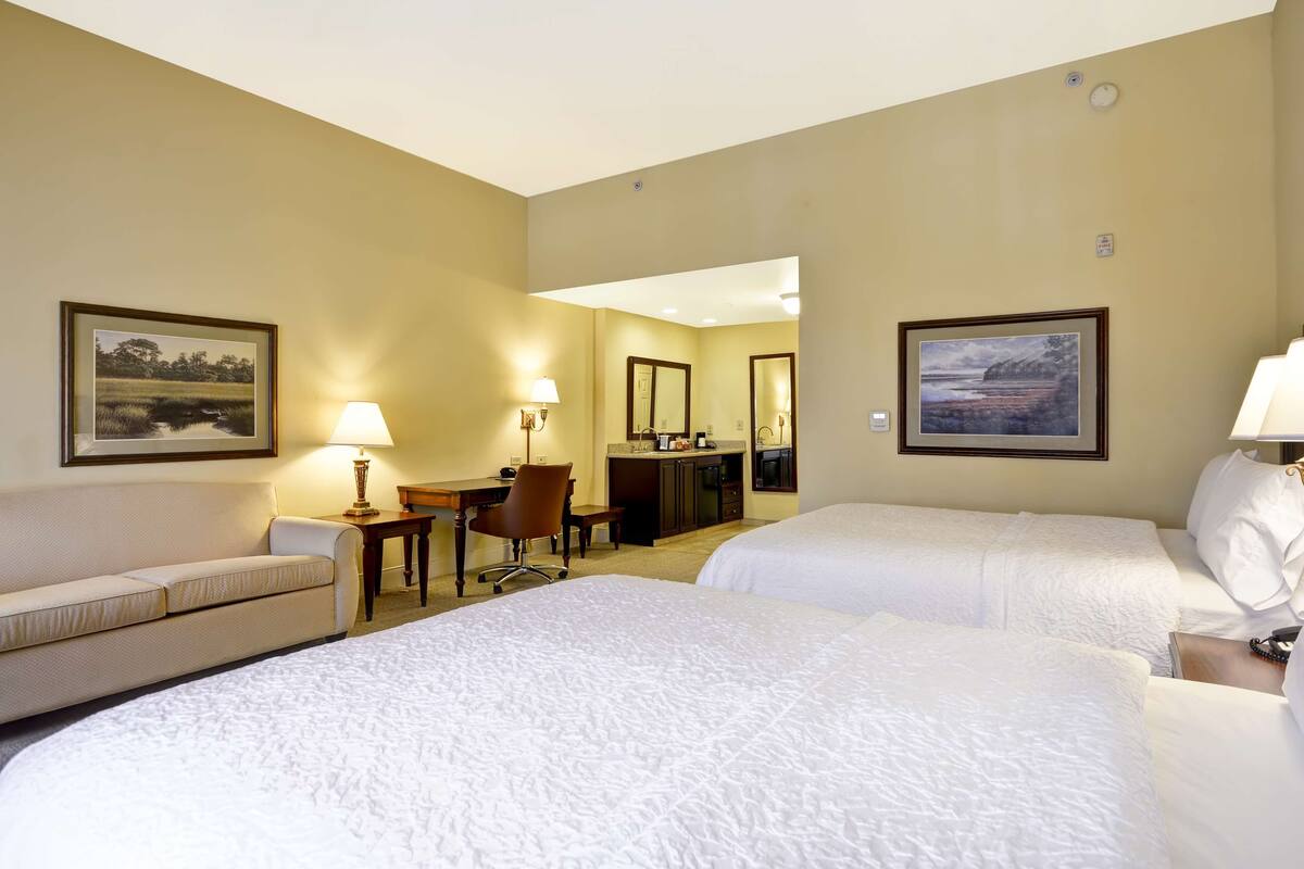 A hotel room with two beds, a desk, a chair, a lamp, and framed pictures on the walls.