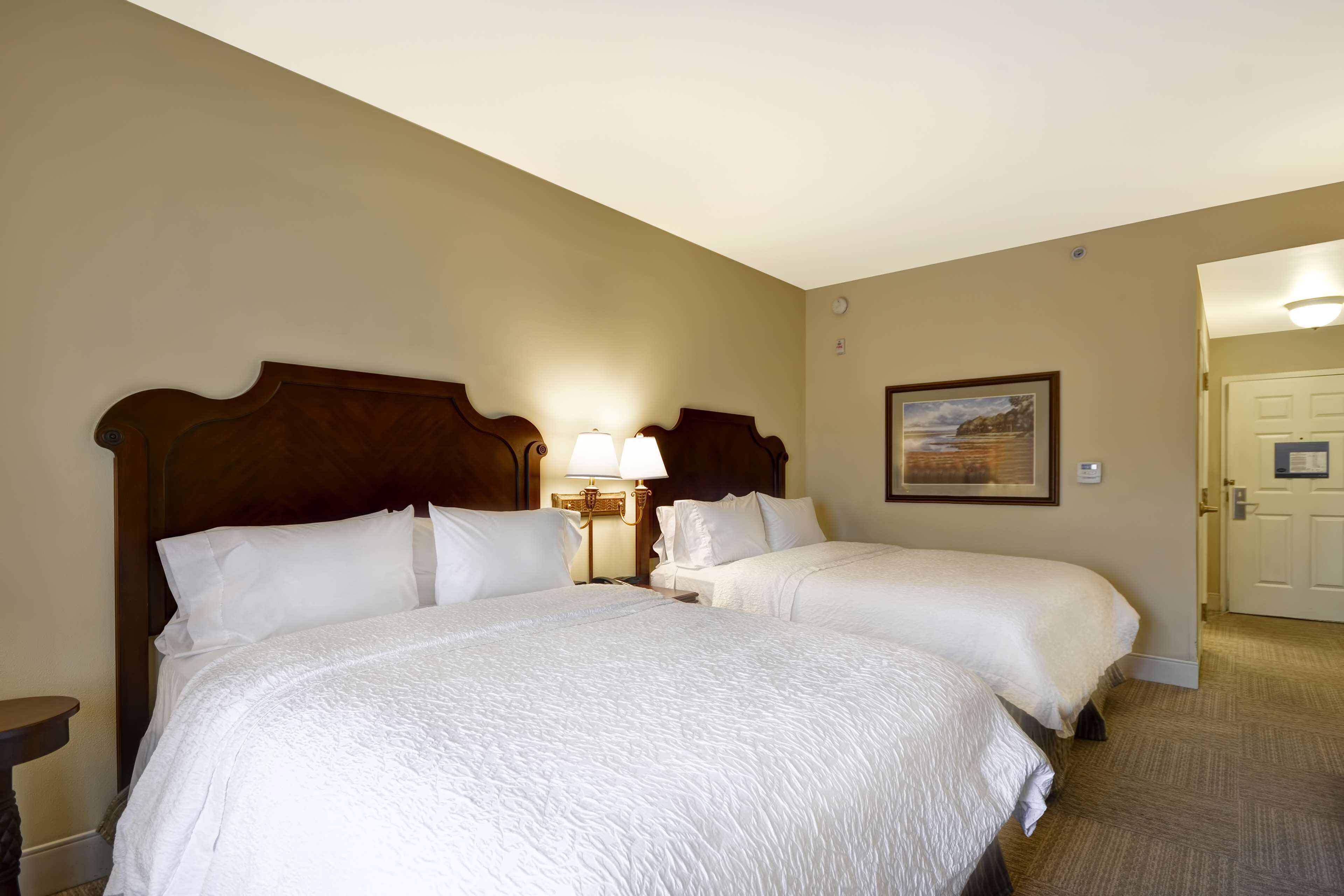 Een hotelkamer met twee bedden, een nachtkastje met een lamp, een lijst aan de muur en een deur aan de rechterkant.