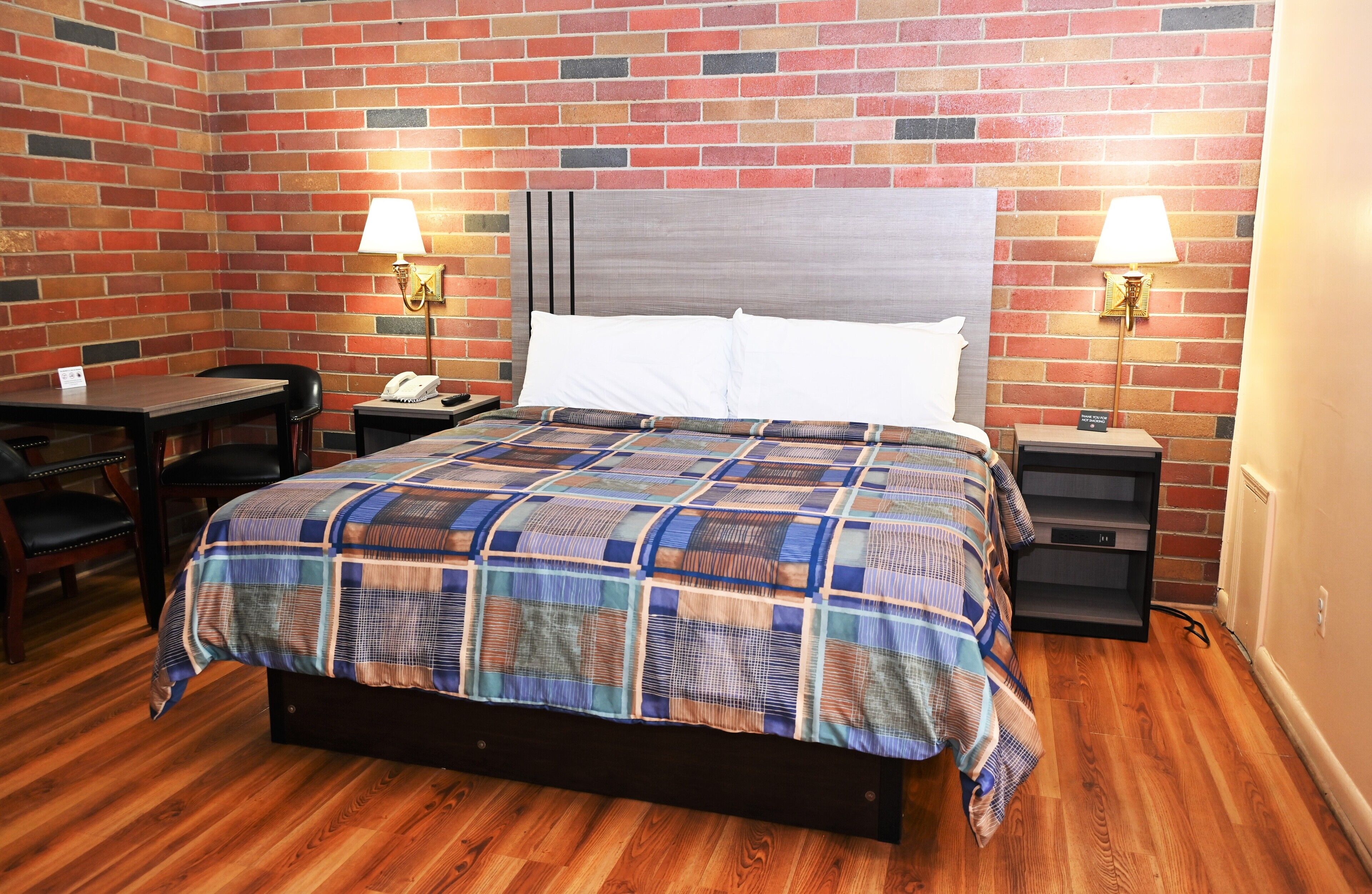 A hotel room with a brick-patterned wall, a wooden floor, a bed with a plaid bedspread, two bedside tables with lamps, a desk, and a chair.