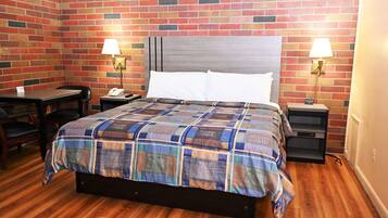 A hotel room with a brick-patterned wall, a wooden floor, a bed with a plaid bedspread, two bedside tables with lamps, a desk, and a chair.