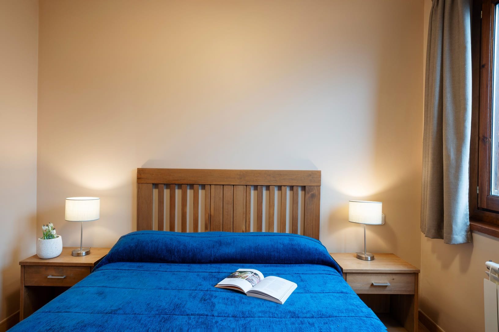 A hotel room with a wooden headboard, a blue bedspread, two bedside lamps, a small potted plant, and a window with curtains.