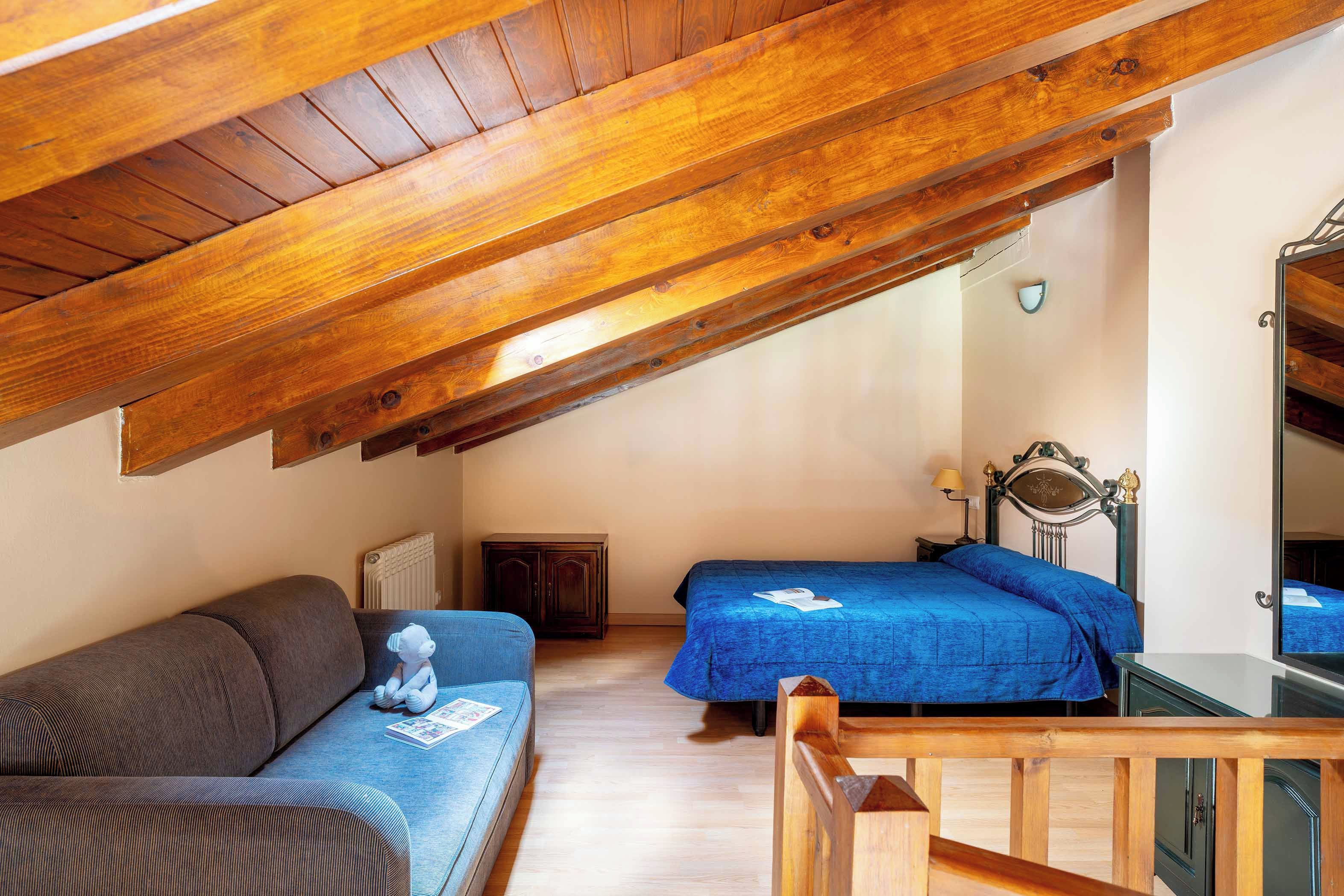A cozy attic bedroom with a wooden ceiling, a sofa, two beds with blue bedding, a dresser, and a mirror.