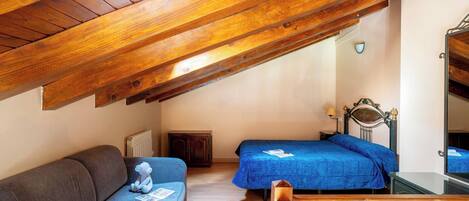 A cozy attic bedroom with a wooden ceiling, a sofa, two beds with blue bedding, a dresser, and a mirror.