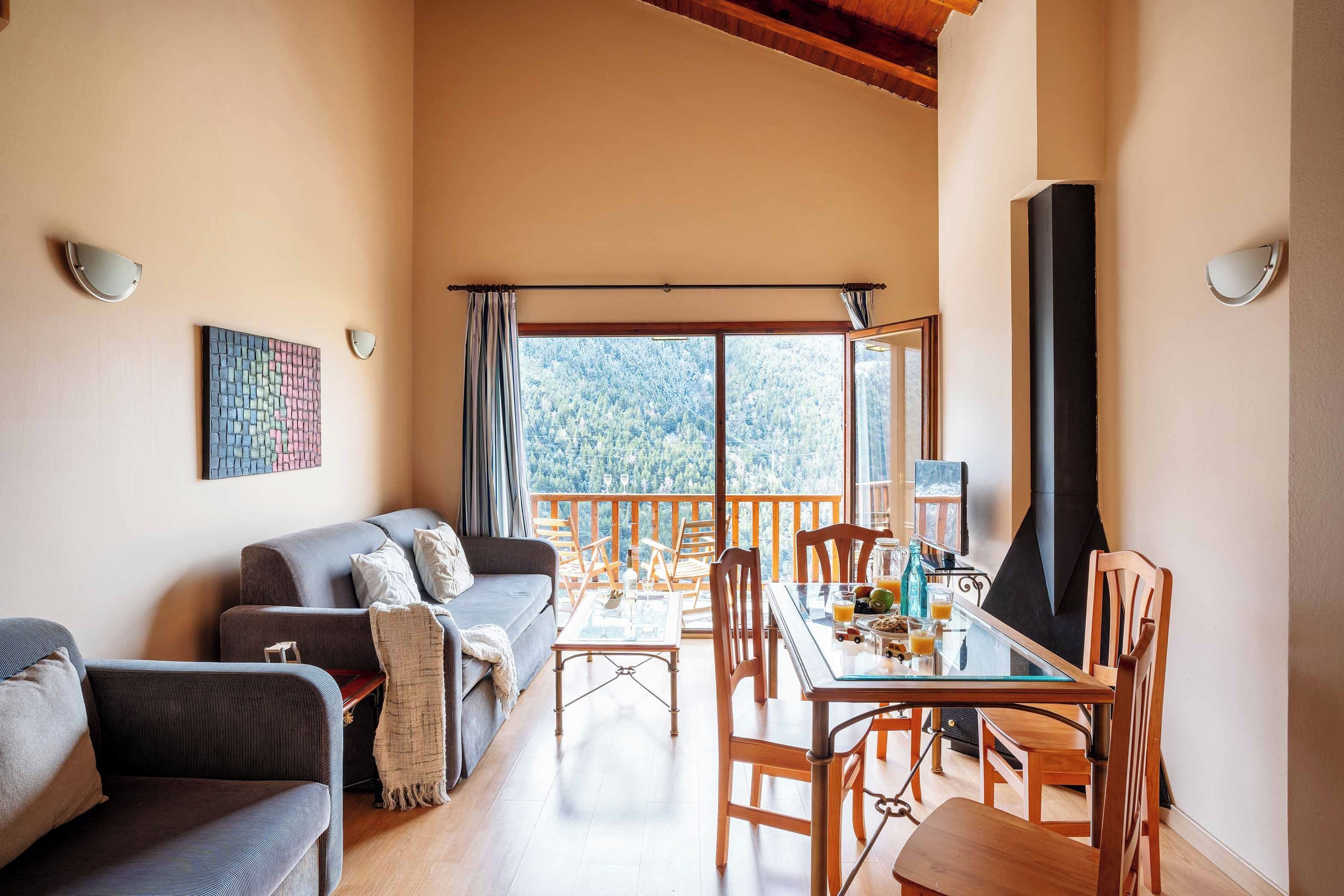 A cozy living room with a sofa, a dining table with chairs, a fireplace, and a large window with a view of trees.