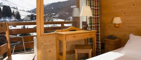 A wooden room with a bed, a desk, and a chair. There is a window with a view of snow-covered mountains.
