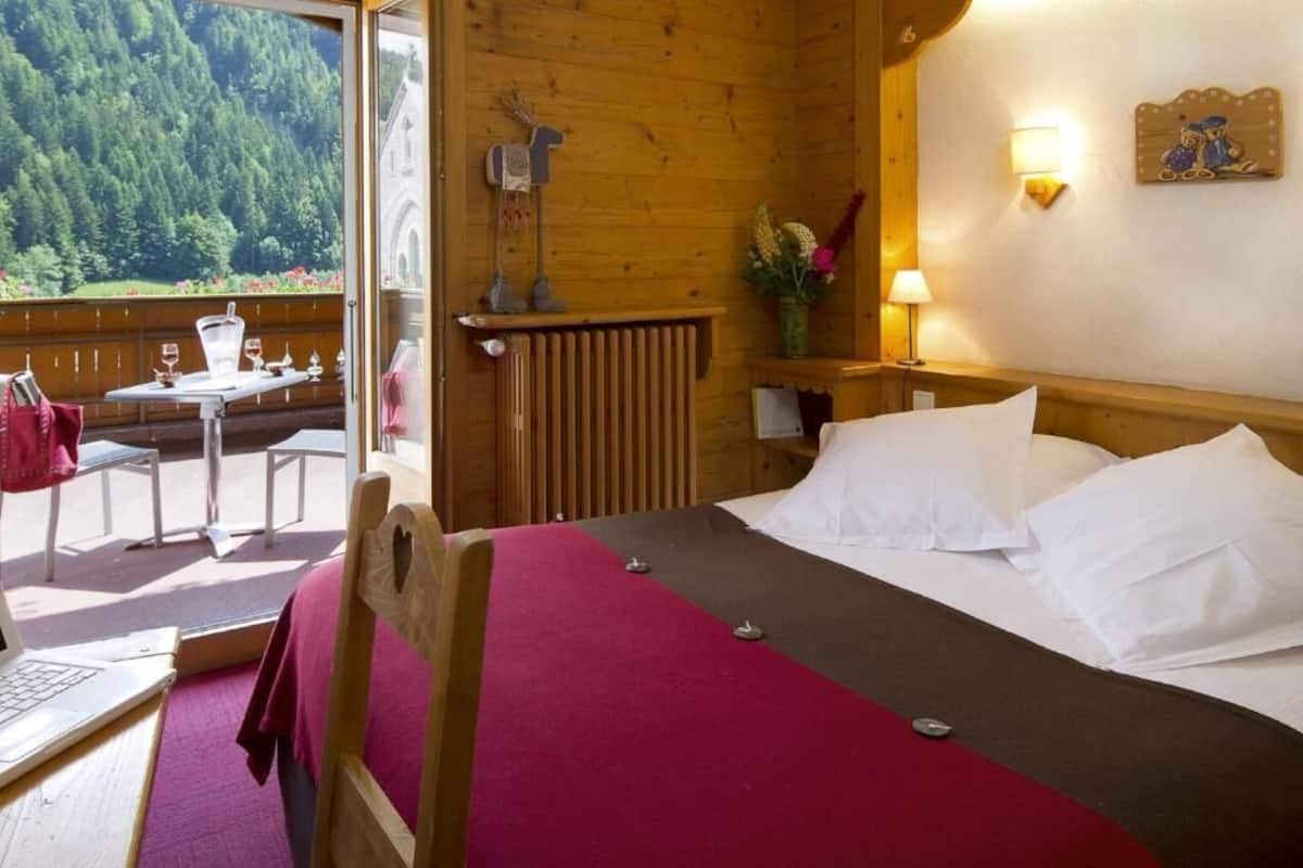 Une pièce en bois avec un lit, une table entourée de chaises, un ordinateur portable et une vue sur une forêt.