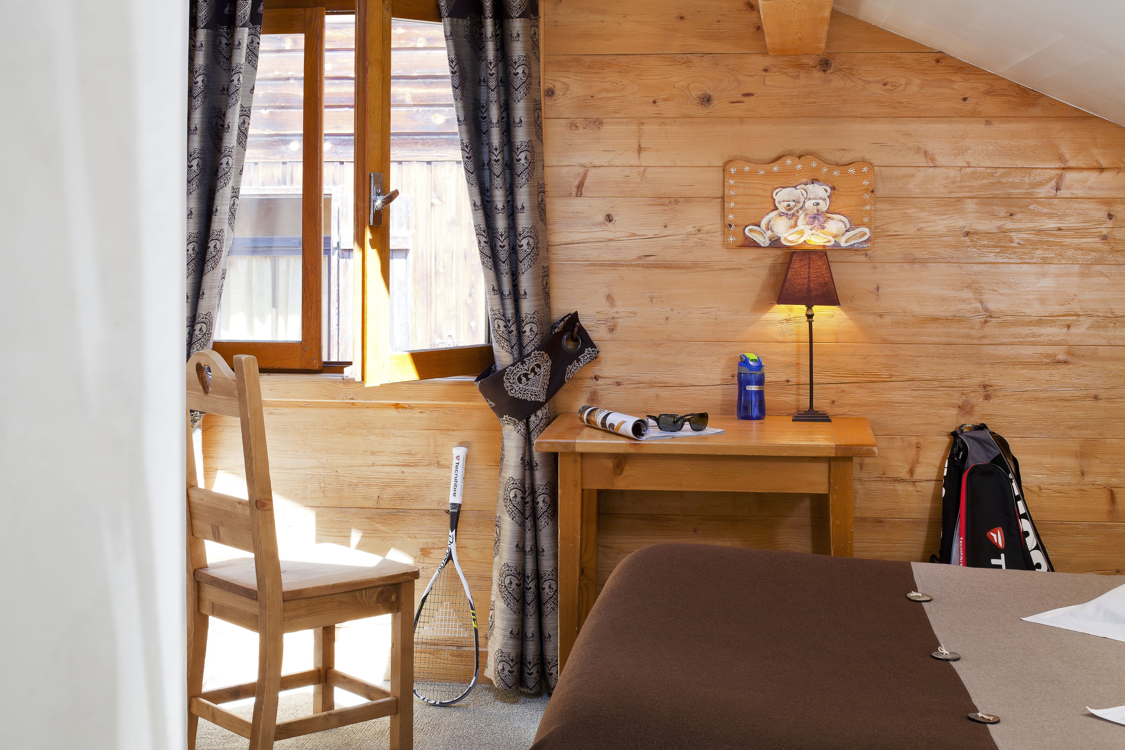 A wooden room with a bed, a desk, a chair, a lamp, a tennis racket, and a window with curtains.