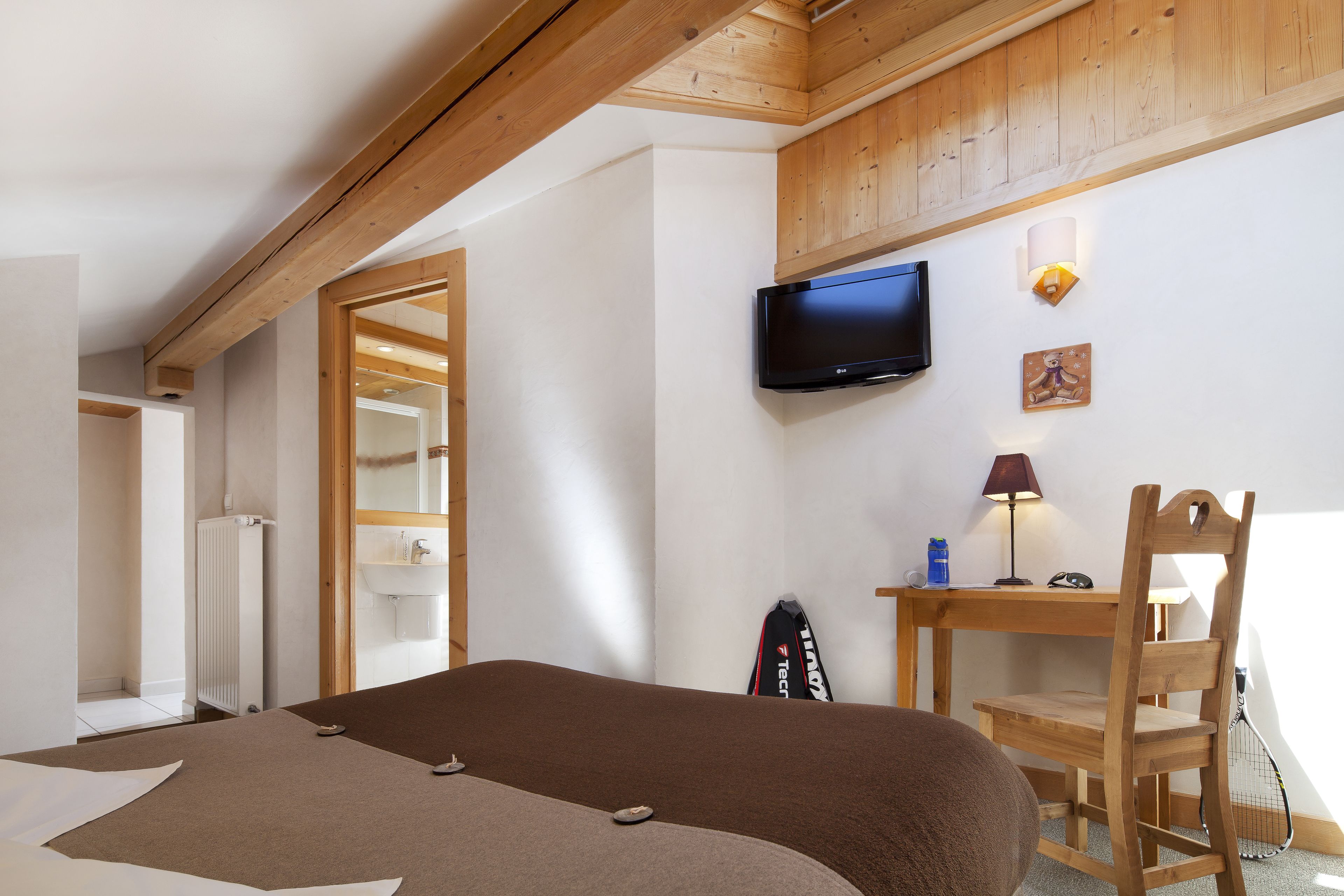 A bedroom with a bed, a TV mounted on the wall, a desk with a chair, and a window.